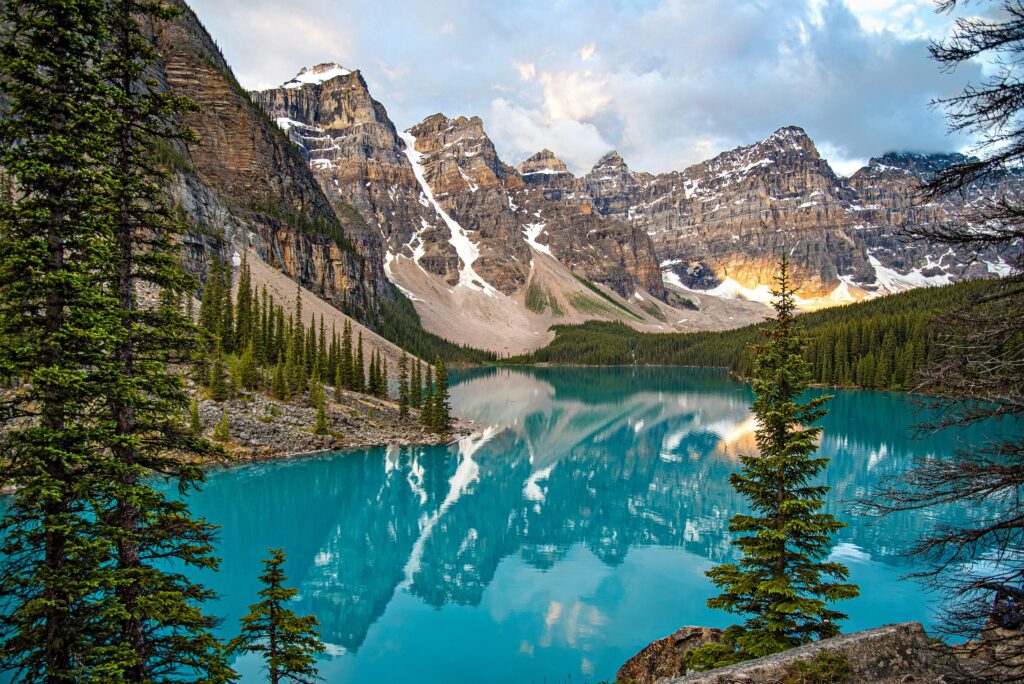 Breathtaking view of Moraine Lake with reflection of snow-covered mountains, pine trees, and vibrant blue water.